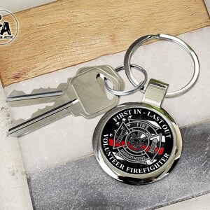 May include: A silver keychain with a black and red round metal charm. The charm has the words "First In - Last Out" and "Volunteer Firefighter" around the edge. The center of the charm has a black and white image of a firefighter helmet and axe with the word "Fire" above it.
