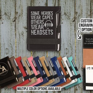 May include: A collection of leatherette notepads in various colors, each with a pen and elastic band. The top notepad is black and features the text "SOME HEROES WEAR CAPES OTHERS WEAR 911 HEADSETS." The image also shows a custom engraving option.