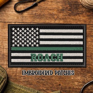 May include: Embroidered patch featuring an American flag design with a green stripe and the name "ROACH". The patch has a black border and is displayed on a wooden surface. The text "EMBROIDERED PATCHES" is below.