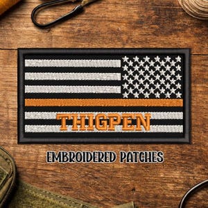 May include: Embroidered patch featuring a stylized American flag design with black and white stripes, a field of white stars, and an orange stripe. The name "THIGPEN" is embroidered in orange. The patch is framed in black.