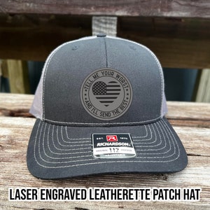 May include: A black and gray trucker hat with a laser-engraved leatherette patch featuring an American flag heart and the text "Tell me your worst and I'll send the best."