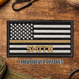 May include: Embroidered patch featuring an American flag design with a gold stripe and the name "SMITH". The patch has a black border and is displayed on a wooden surface. The text "EMBROIDERED PATCHES" is below the patch.
