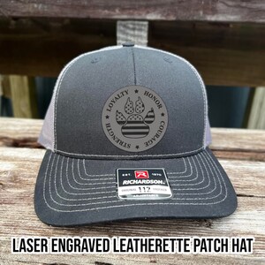 May include: A black and gray trucker hat with a laser engraved leatherette patch featuring an American flag paw print with the words "Loyalty, Honor, Courage, Strength" around it. The hat is adjustable and has a Richardson logo on the back.