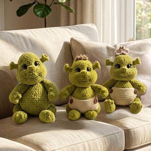 Pode incluir: Três bonecos de crochê feitos à mão com corpos de fio verde, cabeças grandes e orelhas em forma de trompete. Os bonecos têm olhos escuros e sorrisos bordados. Dois dos bonecos usam fraldas de tecido claro com pequenos botões castanhos.