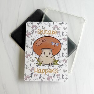 May include: A rectangular tablet case featuring a cartoon shiitake mushroom with a brown cap and a smiling face. The background has a repeating pattern of small mushrooms and dragonflies. The text "Shitake Happens" is printed in brown.