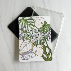 May include: A rectangular tablet case featuring a white background with a pattern of green and white monstera leaves. The text "I should be watering plants" is printed in the center in a dark green script font. A clear protective case is also visible.