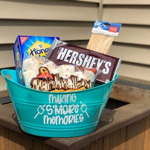 May include: A turquoise metal bucket with the text "Making S'more Memories" on the side. The bucket is filled with s'mores ingredients, including chocolate bars, marshmallows, graham crackers, and wooden skewers.