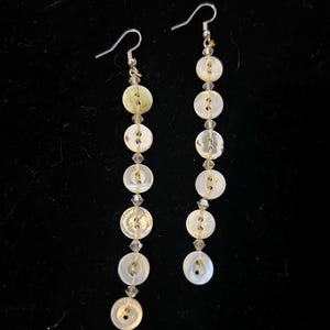 May include: Dangling earrings featuring a series of white, pearlescent buttons strung together with clear beads. The earrings have silver-colored fish hook ear wires and are set against a black background.