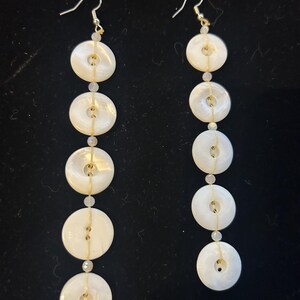 May include: A pair of dangling earrings featuring a series of five white, circular, iridescent shell beads. The beads are strung together with a thin, gold-colored wire and small, clear beads. The earrings have gold-colored ear wires.