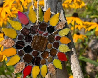 May include: A circular mosaic ornament shaped like a sunflower, featuring a dark gray grout base with inlaid glass pieces in shades of yellow, orange, red, and brown. The piece includes a jute twine loop for hanging from a wooden support structure.