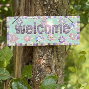 Könnte beinhalten: Ein rechteckiges Mosaikschild mit dem Wort welcome aus dunkelvioletten Fliesen. Der Hintergrund besteht aus hellgrünen Fliesen mit Blumenmustern in Rosa- und Violetttönen sowie schillernden Glasakzenten. Das Schild hängt an einem dünnen schwarzen Draht.