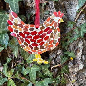 May include: A chicken-shaped mosaic ornament crafted from small, irregular glass pieces in shades of red, orange, and yellow. The mosaic features a white grout base and is suspended by a thin, translucent red ribbon tied in a bow at the top.