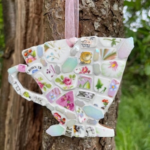 May include: A mosaic teacup ornament made from broken china. The teacup design incorporates floral patterns in pink, yellow, and blue, with a pink ribbon for hanging. The text "CROWN TR" is visible on the side of the cup.