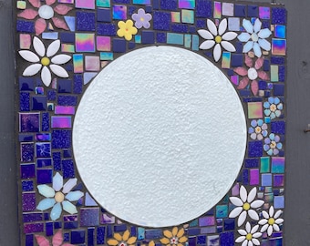 May include: A square mosaic mirror with a round mirror in the center. The mosaic is made of small blue, purple, pink, and yellow tiles. The border of the mosaic is decorated with white and yellow flowers.