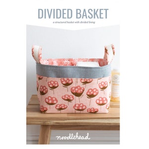 May include: A pink fabric basket with a divided lining and white handles. The basket has a floral pattern with pink flowers and brown stems. The text "DIVIDED BASKET" and "a structured basket with divided lining" is above the basket. The text "noodlehead" is below the basket.