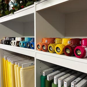 May include: A white shelving unit filled with spools of colorful thread and bolts of fabric. The thread spools are in various colors, including black, white, blue, orange, yellow, and pink. The fabric bolts are yellow and cream-colored.
