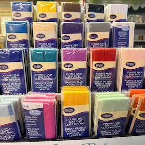 May include: A display of multiple packages of Wrights double fold bias tape quilt binding in various solid colors including blue, yellow, brown, black, white, purple, gray, red, pink, green, and orange, each labeled with 0.875 inches width.