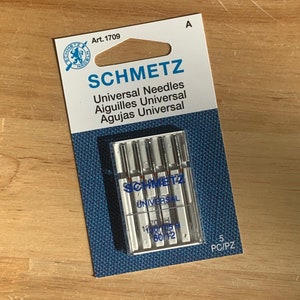 May include: A pack of 5 Schmetz Universal needles in a clear plastic case. The needles are silver and the case is labeled with the Schmetz logo and the text "Universal". The pack is labeled with the text "Art. 1709" and "5 PC/PZ".