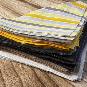 May include: A stack of fabric squares in various colors, including yellow, gray, and black. Some squares have horizontal stripes. The fabric appears to be linen or a similar woven material. The squares are stacked on a wooden surface.