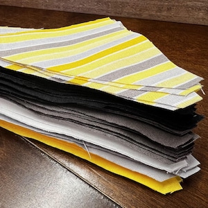 May include: A stack of fabric squares in various colors. The top layer features yellow, gray, and white striped fabric. Below are solid squares in black, gray, white, and yellow. The fabrics are neatly stacked on a wooden surface.