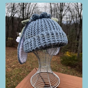 May include: A blue crochet hat with long bunny ears, green accents, and a pom-pom design. The hat is displayed on a wire mannequin head. The image includes the text "PDF Instant Download" and "Crochet".