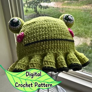 May include: Green crochet frog hat with pink and green embellishments. The hat has a wide brim and is made with a textured stitch. The hat is on a display stand. Digital Crochet Pattern.