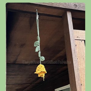 Puede incluir: Una flor de ganchillo amarilla con hojas verdes cuelga de un gancho de metal. El texto "PDF Instant Download" y "Crochet" es visible. También es visible el nombre de la tienda de Etsy "Etsy@AnotherStitchToMake".