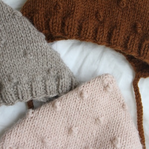 May include: Three knitted baby hats in brown, grey, and pink. The hats have a textured knit pattern and are made with soft yarn.