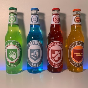 Könnte beinhalten: Vier Glas-Limonadenflaschen mit farbigen Flüssigkeiten und passenden Verschlüssen. Jede Flasche hat ein einzigartiges Etikett mit Text und Grafiken: Speed Cola, Revive Soda, Juggernog Soda und Double Tap Root Beer. Die Flaschen stehen auf einer weißen Oberfläche.