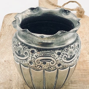 Könnte beinhalten: Eine silberfarbene Vase im antiken Stil mit gerüschtem Rand. Die Vase weist detaillierte, geätzte Blumen- und Scrollmuster um die Mitte und vertikale Linien am unteren Teil auf. Die Vase ist wahrscheinlich aus Metall und hat einen dunklen Innenraum.