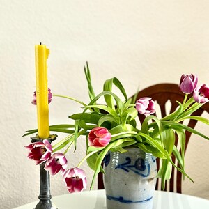 Könnte beinhalten: Ein Stillleben mit einer gelben Kerze in einem Metallkerzenhalter und einer kleinen Keramikvase mit Tulpen. Die Tulpen haben rote und violette Blütenblätter und grüne Blätter. Die Vase hat ein blaues Blumenmuster.