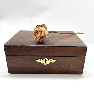 Könnte beinhalten: Eine dunkelbraune Holzkiste mit einem goldfarbenen Schlüssellochdetail. Eine getrocknete Rose mit gelben und roten Blütenblättern liegt auf der Oberseite der Kiste. Die Kiste ist rechteckig und hat ein schlichtes, elegantes Design.