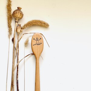 Könnte beinhalten: Ein Holzlöffel mit rundem Kopf und langem Stiel. Auf dem Kopf des Löffels ist eine Cartoonfigur und der Text "Kinder Badebürste" eingraviert. Getrocknete Pflanzen und Samenkapseln sind links vom Löffel angeordnet.