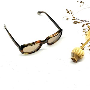 Könnte beinhalten: Rechteckige Brille mit Schildpattmuster und getönten Gläsern. Die Brille ist vor einem weißen Hintergrund platziert, mit einem Trockenblumenarrangement. Der Rahmen weist Brauntöne, Grün und Orange auf.