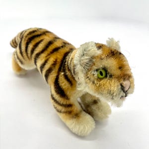 Könnte beinhalten: Ein kleines Tiger-Plüschtier. Es hat einen beigefarbenen Körper mit schwarzen Streifen. Der Tiger hat grüne Augen, eine rosa Nase und weiße Pfoten. Das Spielzeug ist in einer Laufposition dargestellt, mit leicht erhobenem Kopf.