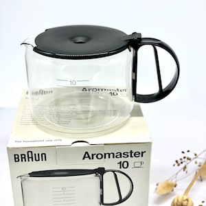 Könnte beinhalten: Eine transparente Braun Aromaster 10-Tassen-Kaffeekaraffe mit schwarzem Deckel und Griff. Die Karaffe steht auf einer weißen Schachtel mit dem Braun-Logo und dem Aufdruck "Aromaster 10". Die Karaffe hat Messmarkierungen.