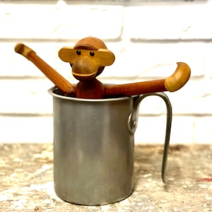 Könnte beinhalten: Ein hölzernes Affenspielzeug mit braunen und braunen Details ist in einem silbernen Metallkrug mit Henkel platziert. Der Affe hat große, runde Augen und ein breites Lächeln.