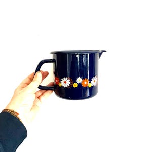 Könnte beinhalten: Ein marineblauer Emaille-Krug mit schwarzem Griff und Ausguss. Der Krug ist mit einem Band aus bunten Gänseblümchen in Weiß, Orange und Gelb verziert. Der Krug wird von einer Person gehalten.