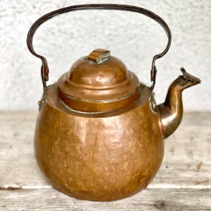 Könnte beinhalten: Ein antiker Kupferkessel mit patinierter Oberfläche und einem dunkelbraunen Henkel. Der Kessel hat einen runden Korpus und einen gewölbten Deckel.
