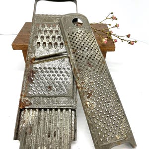 Könnte beinhalten: Vintage Metallreibe mit Griff und separater Reibefläche. Die Reibe hat eine rechteckige Form mit unterschiedlich großen Löchern und einem leicht rostigen Aussehen. Die Reibe wird wahrscheinlich zur Zubereitung von Speisen verwendet.
