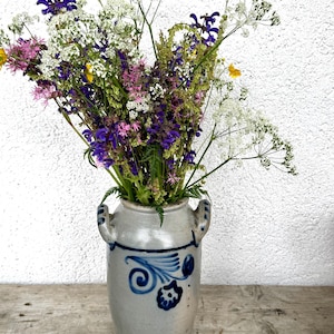 Könnte beinhalten: Ein Strauß Wildblumen in einer Vintage-Vase aus hellgrauer Keramik mit blauen Blumenmustern. Die Blumen umfassen lila, weiße, rosa und gelbe Blüten. Die Vase hat zwei Griffe und steht auf einer Holzoberfläche.