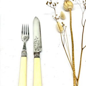 Könnte beinhalten: Ein Vintage-Gabel- und Messerset mit elfenbeinfarbenen Griffen und aufwendigen Silberdetails. Die Messerklinge und die Gabelzinken sind silberfarben. Das Set wird auf einer weißen Oberfläche mit getrockneten floralen Akzenten präsentiert.