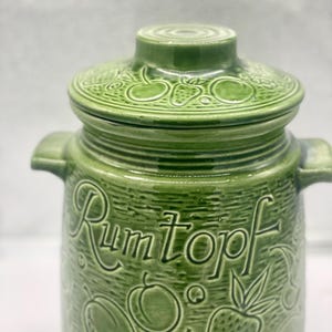 Könnte beinhalten: Grüner Keramik-Rumtopf-Topf mit Deckel. Die Dose hat geprägte Fruchtdesigns und das Wort "Rumtopf". Der Deckel hat auch Fruchtdesigns. Die Dose hat zwei Griffe an den Seiten.