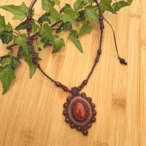 Può includere: Una collana macramè fatta a mano con un grande ciondolo ovale in pietra rosso-arancio. Il ciondolo è circondato da un elaborato design macramè con piccole perline rotonde marrone-rossastre. La collana è marrone e poggia su una superficie di legno.
