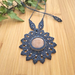Op de afbeelding: Een handgemaakte marineblauwe macramé ketting met een grote, ronde, lichtbruine stenen hanger. De hanger is omgeven door ingewikkelde macramé details en kleine zilveren kralen. De ketting wordt gepresenteerd op een houten oppervlak.