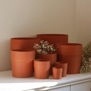 Peut inclure: Une collection de six pots de fleurs cylindriques de couleur terre cuite de tailles variées disposés en groupe. Les pots présentent une finition lisse et mate avec un design simple à parois droites et une fine arête horizontale près du bord.