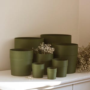Peut inclure: Un ensemble de six pots de fleurs cylindriques vert olive de tailles variées disposés en groupe. Les pots présentent une finition mate lisse avec une fine nervure horizontale près du bord supérieur. Un pot contient un bouquet de fleurs séchées.