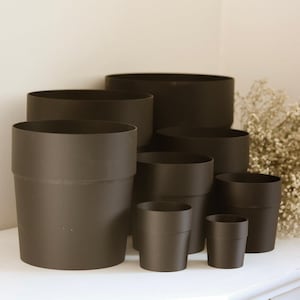 May include: A collection of seven matte black cylindrical plant pots of varying sizes arranged in a cluster. Each pot features a smooth, solid finish with a simple, straight-sided design and a slightly wider rim at the top edge of the container.
