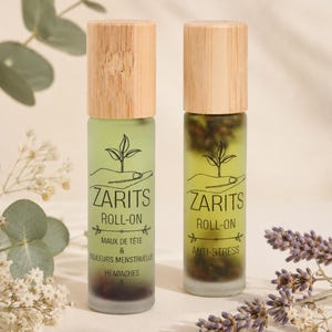 Peut inclure: Deux flacons roll-on en verre cylindriques avec des bouchons en bois clair. Le flacon de gauche est en verre dépoli vert avec le texte ZARITS ROLL-ON MAUX DE TÊTE & DOULEURS MENSTRUELLES, celui de droite est transparent avec ZARITS ROLL-ON ANTI-STRESS.