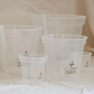 May include: A collection of translucent plastic plant pots in various sizes, each featuring a black printed logo of a hand holding a small sprout with the text ZARITS underneath. The pots have a wide rim and a tapered cylindrical shape.
